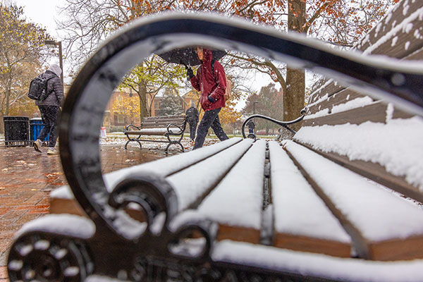 A student walking in the snow with an umbrella is framed through the armrest of a snowcovered bench on campus